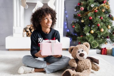 Gülümseyen Afrikalı Amerikalı kız, hediye kutusu ve oyuncak ayıyla yerde oturmuş kameraya bakıyor. 