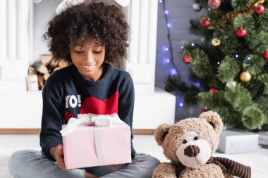 Neşeli Afro-Amerikalı kız elinde Noel hediyesi tutarken peluş ayının yanında otururken bulanık arka planda