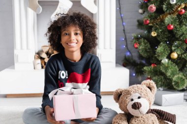 Mutlu Afro-Amerikan kızı elinde hediye tutarken kameraya bakıyor. Arka planda bulanık bir şömine ve Noel ağacı var.