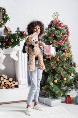 Mutlu Afrikalı Amerikalı kız elinde hediye kutusu ve oyuncak ayıyla süslenmiş Noel ağacı ve şöminenin yanında.