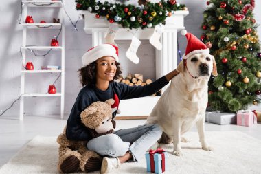 Gülümseyen Afrikalı Amerikalı kız oyuncak ayıyla yerde otururken Labrador köpeğini okşuyor. 
