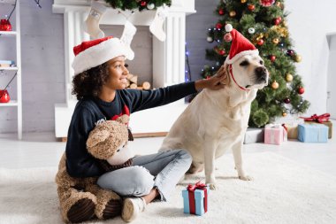 Neşeli Afrikalı Amerikalı kız, bulanık arka planda oyuncak ayıyla yerde otururken Labrador köpeğini okşuyor.