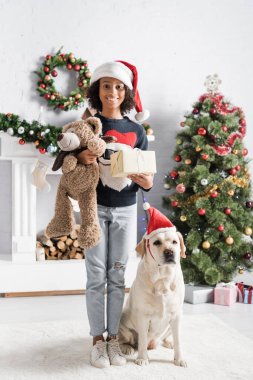Neşeli Afrikalı Amerikalı kız hediye kutusu ve labrador köpeği ve noel ağacının yanında oyuncak ayı tutuyor. 
