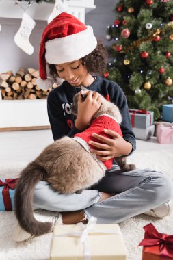 Gülümseyen Afrikalı Amerikalı kız, Noel Baba şapkalı, pofuduk kediyi kucaklarken hediye kutularının ve bulanık arka planda Noel ağacının yanında oturuyor.