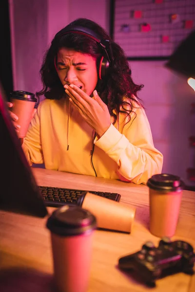 厌倦了的非洲裔美国游戏玩家一边打呵欠一边拿着咖啡走近电脑和操纵杆 模糊了前景 — 图库照片