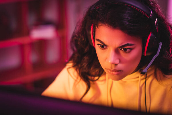 Focused african american player in headset looking at computer monitor on blurred foreground 