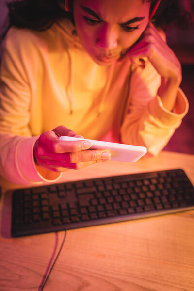 Pensive african american gamer using smartphone near computer keyboard on blurred background 