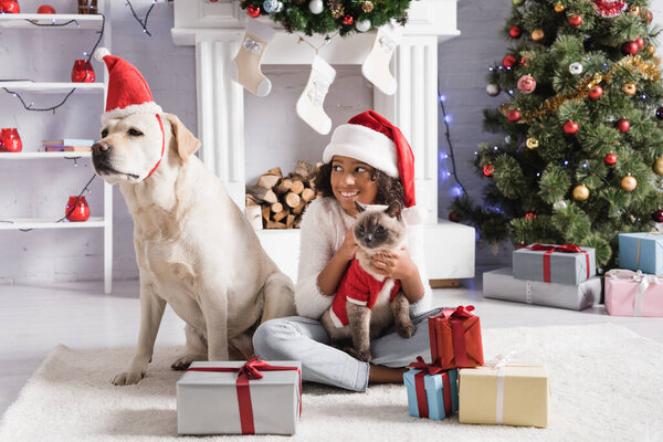 happy african american girl holding cat while sitting near gift boxes and looking at labrador dog  
