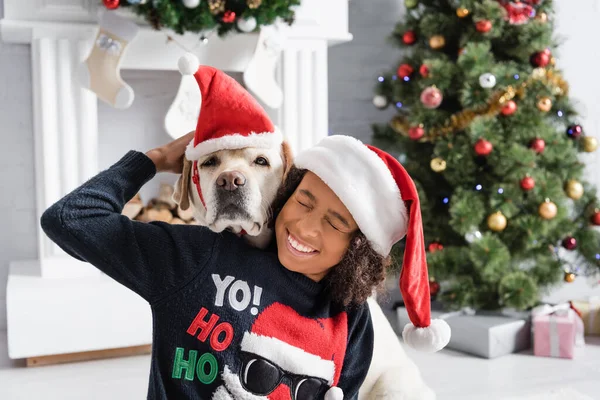 Excited african american girl with closed eyes hugging labrador dog near christmas tree on blurred background — Stock Image Excited African American Girl Closed Eyes Hugging Labrador Dog Christmas Stock Picture