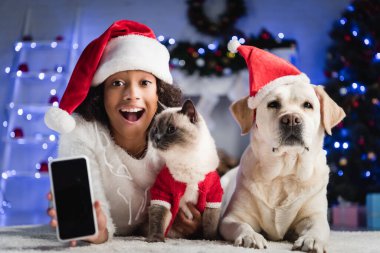 Beyaz ekranlı akıllı telefon gösteren Afro-Amerikalı bir kız yerde bulanık arka planda kedi ve köpeğin yanında yatıyor.  