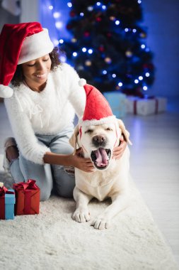 Gülümseyen Afrikalı Amerikalı kız esneyen av köpeğine Noel Baba şapkası takıyor, bulanık arkaplanda hediyelerin yanında oturuyor.