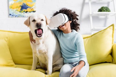 Kıvırcık Afrikalı Amerikalı kız, VR kulaklıklı, esneyen köpeği kucaklayan, bulanık arka planda koltukta oturan.
