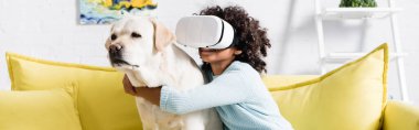 VR kulaklıklı Afrikalı Amerikalı kız evdeki sarı kanepede otururken köpeğe sarılıyor.