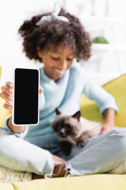Beyaz ekran ile akıllı telefon gösteren Afrikalı Amerikalı bir kız, kediye bakıyor ve bulanık arka planda koltukta oturuyor.