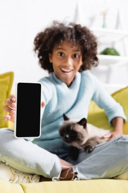 Beyaz ekranlı akıllı telefon gösteren, kediyi okşayan ve arka planda bulanık koltukta oturan mutlu Afrikalı Amerikalı kız.