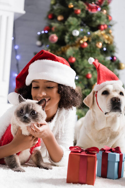 happy african american girl in santa hat cuddling cat while lying on floor near labrador dog on blurred background