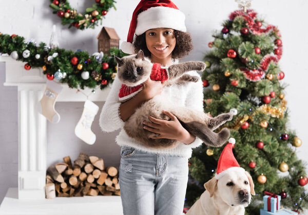 happy african american girl holding furry cat near labrador dog, decorated fireplace and christmas tree on blurred background