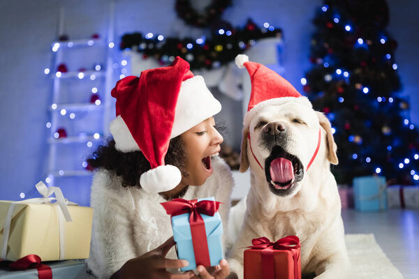 african american girl and labrador in santa hat yawning while lying on floor near gift boxes on blurred background