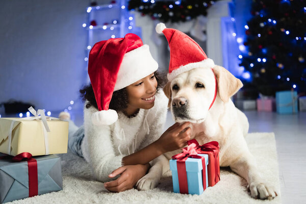 cheerful african american girl stroking labrador dog while lying on floor near gift boxes on blurred background