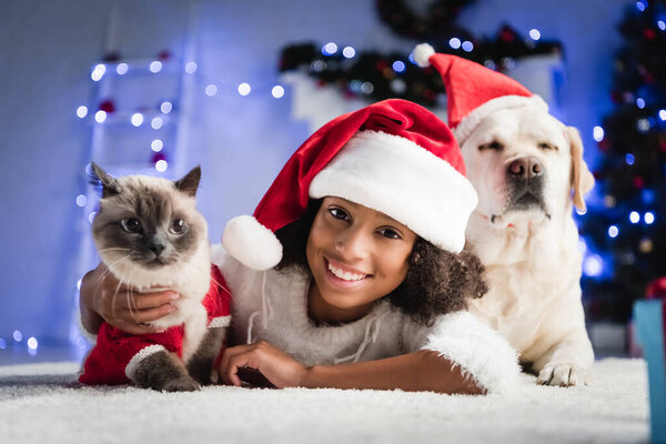 happy african american girl in santa hat lying on floor near labrador and cat on blurred background