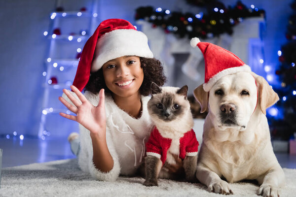Happy african american girl with waving hand lying on floor near cat and labrador in santa hat on blurred background