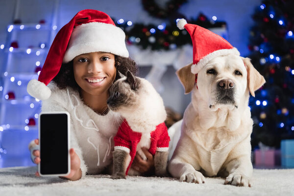 Smiling girl showing smartphone with blank screen and lying on floor near cat and retriever on blurred background 