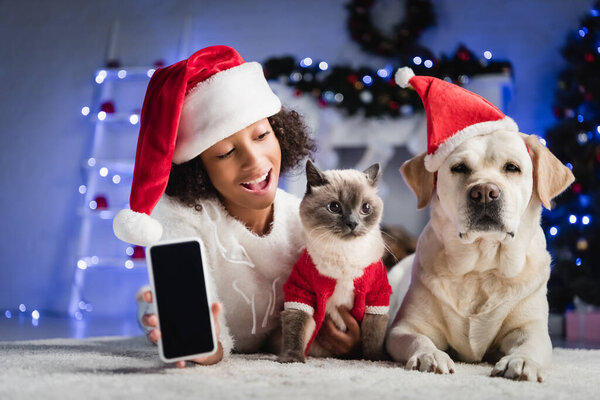 Excited african american girl showing smartphone with blank screen and looking at cat, while lying on floor on blurred background  