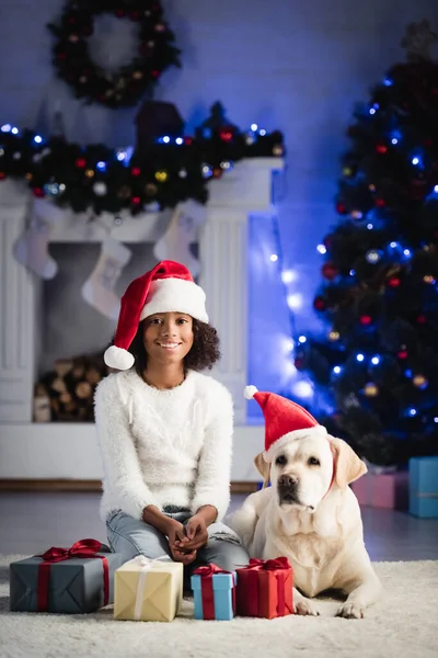 Labrador Mit Weihnachtsmütze Neben Lächelndem Afrikanisch Amerikanischem Mädchen Sitzend Der — Stockfoto