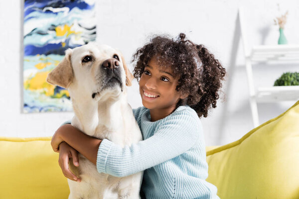 Smiling african american girl hugging and looking at labrador, while sitting on yellow sofa at home, on blurred background