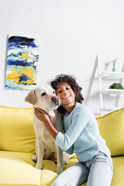 Curly african american girl embracing retriever, while sitting on yellow sofa at home, on blurred background