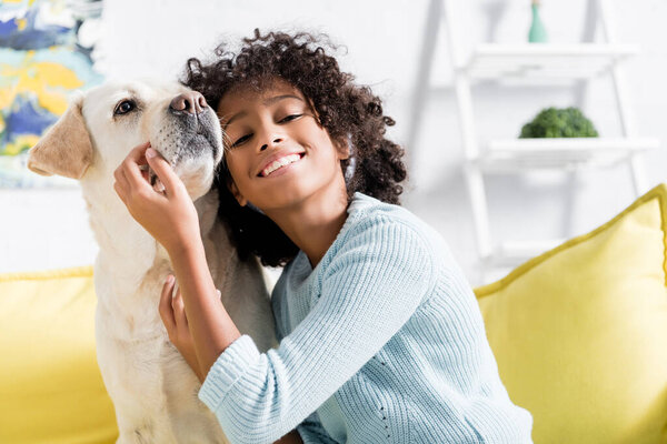 Smiling african american girl leaning on retriever, while sitting on yellow sofa at home, on blurred background 