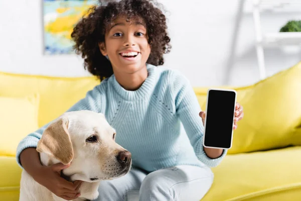 快乐的非洲裔美国女孩 一边在家里抱着拉布拉多 一边在模糊的背景下展示着带有空白屏幕的手机 — 图库照片