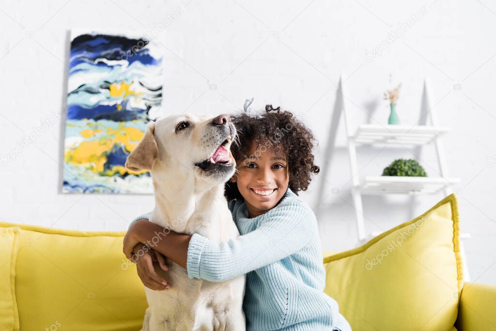 Happy curly girl embracing labrador barking, while sitting on yellow sofa at home