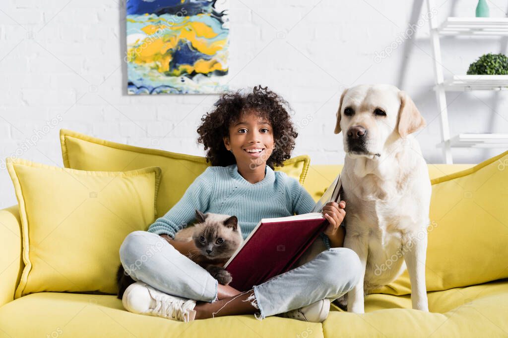 Happy curly african american girl with open book, embracing cat, while sitting near retriever on sofa at home