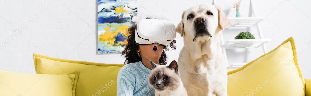 Curly african american girl with open mouth wearing vr headset and sitting near retriever and cat on sofa at home, banner