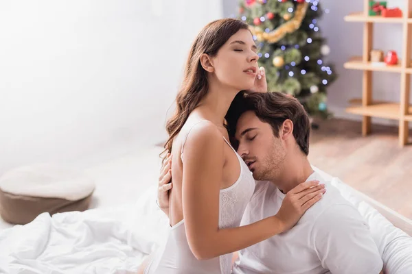 紧闭双眼的充满热情的男人与迷人的年轻女子拥抱在被装饰过的圣诞树旁 背景模糊 — 图库照片