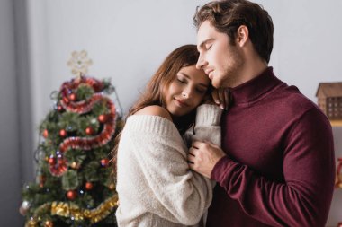 Sıcak kazaklı bir çift, arka planda süslenmiş Noel ağacının yanında sarılıyor.