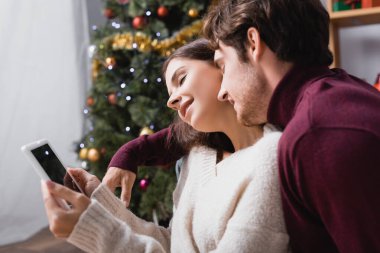 Kazaklı kadın elinde dijital tabletle erkek arkadaşının yanında boş ekran ve bulanık arka planda noel ağacı tutuyor.