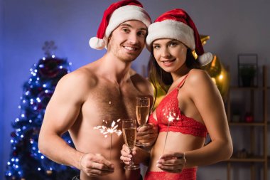Noel Baba şapkalı mutlu ve seksi çift ellerinde şampanya bardakları ve bulanık ışıkların yanında maytap tutuyorlar.