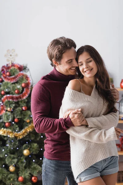 Casal Satisfeito Suéteres Quentes Abraçando Perto Árvore Natal Decorada Fundo — Fotografia de Stock