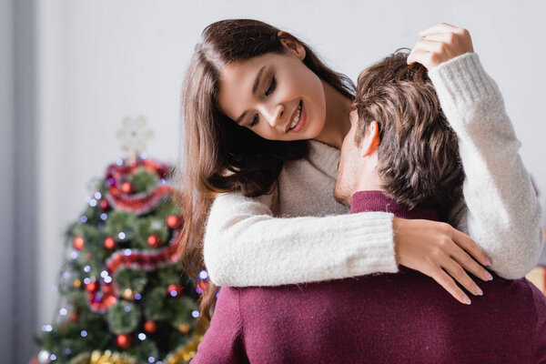 passionate man kissing happy young woman in sweater near christmas tree on blurred background