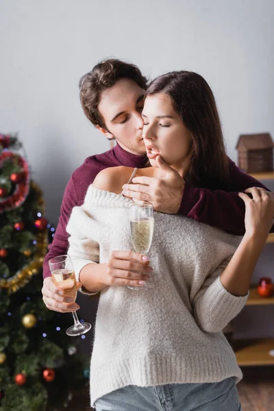 情熱的な男は色気のある女性の唇に触れながらガラスを持っていますシャンパンとクリスマスツリーの近くにぼやけた背景に — ストック写真