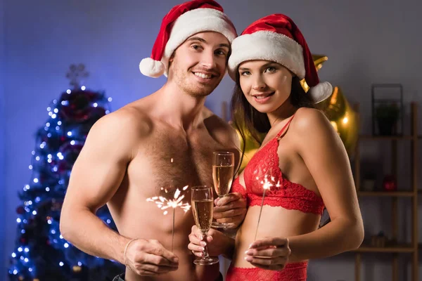 happy and sexy couple in santa hats holding glasses with champagne and sparklers near blurred lighting