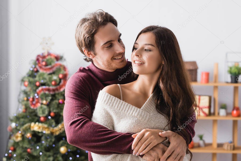 happy couple in warm sweaters hugging near decorated christmas tree on blurred background