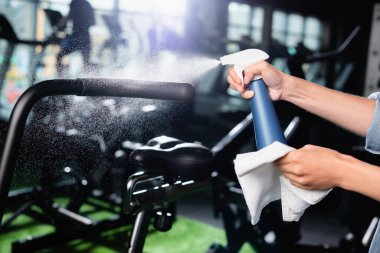 Spor salonundaki egzersiz makinesini temizlerken temizlikçi kadının deterjan sıkarkenki görüntüsü.