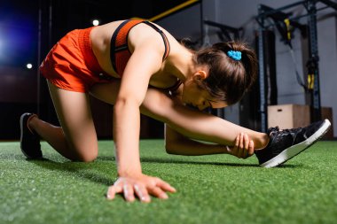 Spor kadını kafasıyla bacağına uzanırken spor salonunda ön planda bulanık bir şekilde geriniyor.
