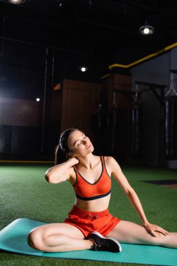 Genç sporcu kadın kapalı gözlerle spor minderinde otururken boynunu ısıtıyor.