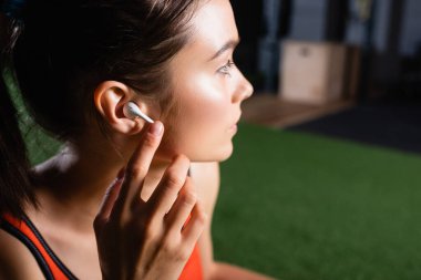 young sportswoman touching wireless earphone and looking away in sports center clipart