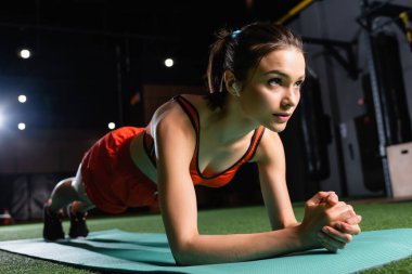 Kulaklıklı genç sporcu kadın spor paspası üzerinde kalas egzersizi yapıyor, geçmişi bulanık.