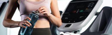 cropped view of sportswoman in bra opening sports bottle while standing on treadmill, banner clipart